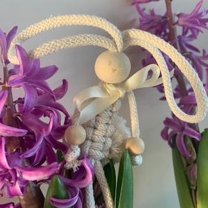 Può includere: Un coniglio in macramé bianco con dettagli in legno e un nastro di raso bianco. Il coniglio è appeso a una pianta di giacinto.