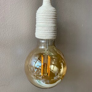 Op de afbeelding: Een decoratieve gloeilamp met een warme, amberkleurige gloed. De lamp heeft een ronde vorm en een witte, met touw omwikkelde basis. De lamp hangt tegen een neutrale grijze muur.