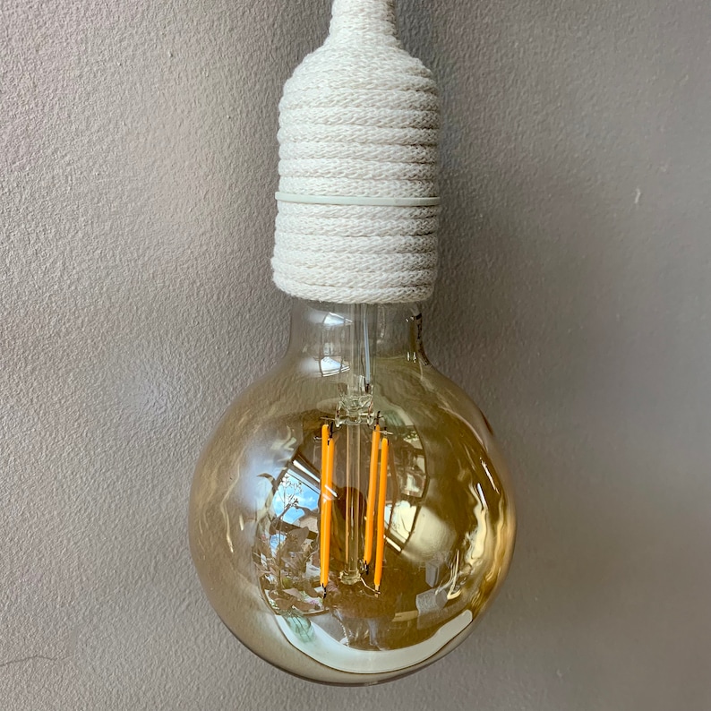 Op de afbeelding: Een decoratieve gloeilamp met een warme, amberkleurige gloed. De lamp heeft een ronde vorm en een witte, met touw omwikkelde basis. De lamp hangt tegen een neutrale grijze muur.