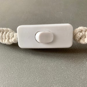 Op de afbeelding: Een close-up van een witte, rechthoekige lichtschakelaar met een afgeronde schakelaar. De schakelaar is aan weerszijden verbonden met cr&egrave;mekleurige macram&eacute; koorden. De achtergrond is een effen grijs oppervlak.