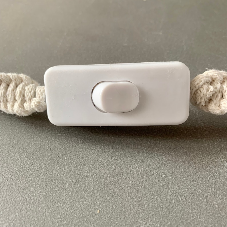 Op de afbeelding: Een close-up van een witte, rechthoekige lichtschakelaar met een afgeronde schakelaar. De schakelaar is aan weerszijden verbonden met cr&egrave;mekleurige macram&eacute; koorden. De achtergrond is een effen grijs oppervlak.