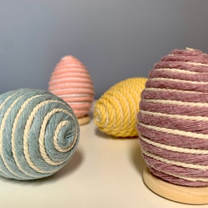 Op de afbeelding: Vier decoratieve paaseieren omwikkeld met touw. De eieren zijn in tinten roze, geel, blauw en paars.