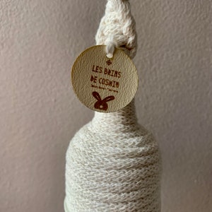 Op de afbeelding: Een decoratieve fles gewikkeld in cr&egrave;mekleurig garen, met een geknoopt bovenstuk en een rond label. Het label bevat de tekst "LES BAINS DE COSHIN" en een konijnenlogo. De fles is ontworpen voor decoratieve doeleinden.