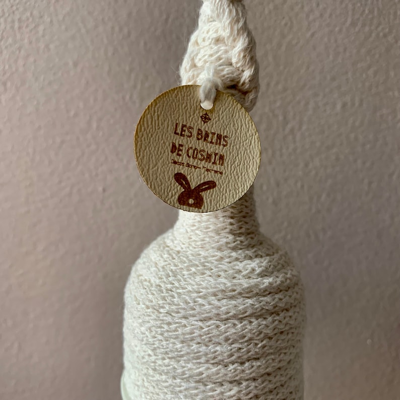 Op de afbeelding: Een decoratieve fles gewikkeld in cr&egrave;mekleurig garen, met een geknoopt bovenstuk en een rond label. Het label bevat de tekst "LES BAINS DE COSHIN" en een konijnenlogo. De fles is ontworpen voor decoratieve doeleinden.
