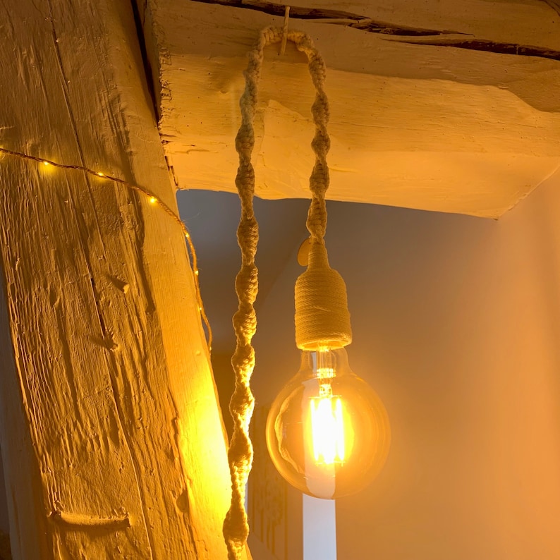 Op de afbeelding: Een hanglamp met een grote, ronde gloeilamp die warm licht uitstraalt. De lamp wordt opgehangen door een gedraaid, cr&egrave;mekleurig touw, bevestigd aan een houten balk. De lamp geeft een warme sfeer.
