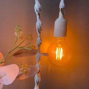 Op de afbeelding: Een close-up van een hanglamp met een warme, oranje gloed. De gloeilamp is een grote, ronde Edison-stijl lamp, omhuld door een cr&egrave;mekleurige macram&eacute;-stijl lampenkap. Een decoratieve plant met roze bladeren staat op de voorgrond.