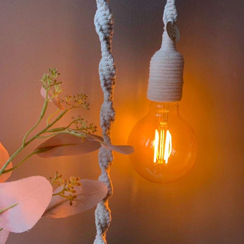 Op de afbeelding: Een close-up van een hanglamp met een warme, oranje gloed. De gloeilamp is een grote, ronde Edison-stijl lamp, omhuld door een cr&egrave;mekleurige macram&eacute;-stijl lampenkap. Een decoratieve plant met roze bladeren staat op de voorgrond.