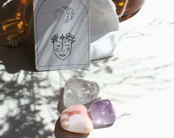 Könnte beinhalten: Ein weißer Kordelzugbeutel und eine Karte mit einem Sonnen- und Monddesign sowie einem Gesicht mit Blumenkranz. Drei polierte Edelsteine: klarer Quarz, rosa Achat und lila Amethyst, werden auf einer weißen Oberfläche gezeigt.