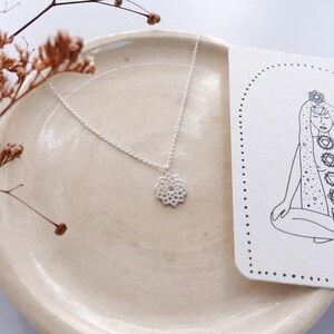 May include: A silver necklace with a floral pendant rests on a beige plate, next to a card with a line drawing of a meditating figure and chakra symbols. Dried brown stems add a natural touch to the composition. The necklace is delicate and elegant.