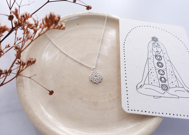 May include: A silver necklace with a floral pendant rests on a beige plate, next to a card with a line drawing of a meditating figure and chakra symbols. Dried brown stems add a natural touch to the composition. The necklace is delicate and elegant.