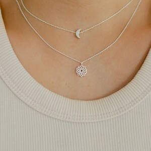 May include: A layered silver necklace set. The top chain features a crescent moon pendant, while the lower chain showcases a floral design. The necklaces are worn over a ribbed, light beige tank top.