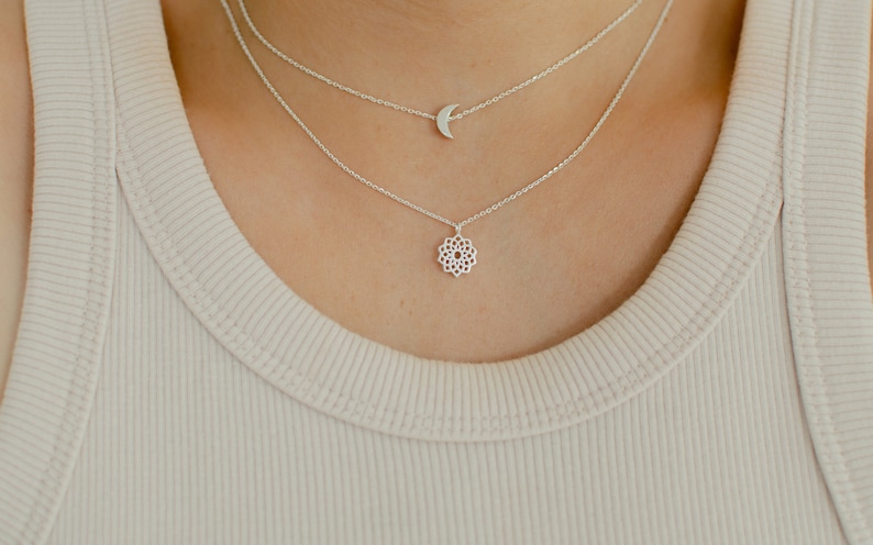 May include: A layered silver necklace set. The top chain features a crescent moon pendant, while the lower chain showcases a floral design. The necklaces are worn over a ribbed, light beige tank top.