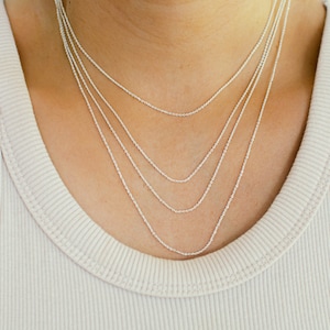 May include: A close-up of a person wearing a beige ribbed tank top. Four delicate silver necklaces of varying lengths are layered around the neck, creating a stylish and elegant look. The necklaces are simple and minimalist.