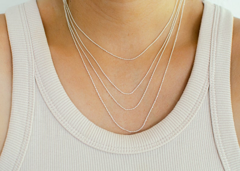 May include: A close-up of a person wearing a beige ribbed tank top. Four delicate silver necklaces of varying lengths are layered around the neck, creating a stylish and elegant look. The necklaces are simple and minimalist.
