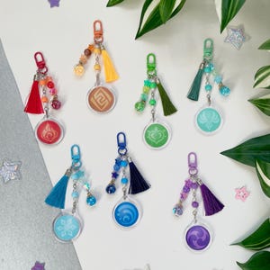 Op de afbeelding: Zeven kleurrijke sleutelhangers met bedels en kwastjes. Elke sleutelhanger heeft een ander elementsymbool: vuur, aarde, wind, water, ijs, bliksem en een hart. De bedels zijn gemaakt van kralen, sterren en kleine ballen.