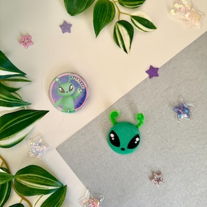 May include: A collection of alien-themed decorations and embellishments. Features a green alien face with black eyes and antennae, a button with an alien graphic and the word "Hallo!", and star-shaped sequins. Green and white leafy plants are also present.