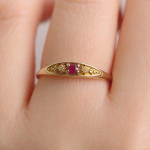 May include: A gold ring featuring a central oval ruby gemstone, flanked by smaller, clear gemstones. The ring has an engraved floral design on the band. The ring is shown on a finger.