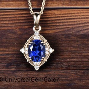 May include: A gold-toned pendant necklace with a large, oval, deep blue gemstone at its center. The gemstone is surrounded by smaller, clear stones and a decorative gold frame. The necklace hangs on a delicate gold chain. The text "UniversalGemGalor" is at the bottom.