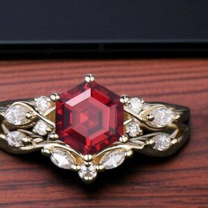 Peut inclure: Un ensemble de bagues en or avec une grosse pierre rouge foncé hexagonale en son centre. La monture est ornée de plus petites pierres précieuses claires et d'éléments décoratifs en or. L'ensemble est posé sur un fond en bois.