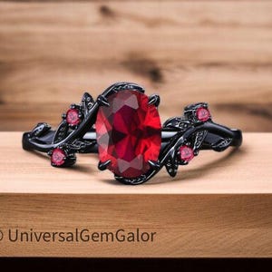 May include: A black ring with a large oval-cut red gemstone as the centerpiece. The ring features a floral design with smaller red gemstones and is set against a wooden background.