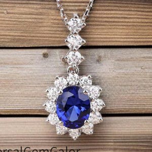 May include: A silver necklace with a large, oval, deep blue gemstone pendant surrounded by smaller white gemstones. The pendant hangs from a delicate chain with three additional white gemstones. The background is a wooden surface.