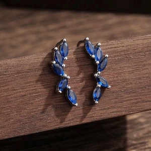Pode incluir: Um par de brincos prateados com um design em forma de folha. Cada brinco apresenta múltiplas pedras preciosas azuis escuras com corte marquesa. Os brincos são colocados sobre uma superfície de madeira escura, realçando o azul vibrante das pedras.