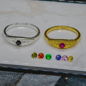 May include: Two rings, one silver and one gold, each with a central gemstone. The silver ring has a black gemstone, while the gold ring features a pink gemstone. Several loose, colorful gemstones are also displayed.