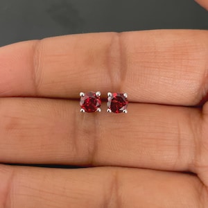 Könnte beinhalten: Ein Paar Ohrstecker mit runden, tiefroten Edelsteinen, die in Silbermetall gefasst sind. Die Ohrringe werden vor einem neutralen Hintergrund gezeigt, der ihre leuchtende Farbe und ihr schlichtes Design hervorhebt. Diese Ohrringe sind ein klassisches Schmuckstück.