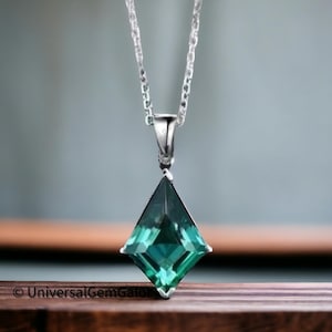 Puede incluir: Un collar de plata con un colgante geométrico verde esmeralda. El colgante tiene forma de diamante con múltiples facetas, que reflejan la luz. La cadena es delicada y el colgante cuelga en el centro. El collar se muestra sobre un fondo borroso.