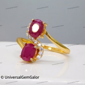 May include: A gold ring with two ruby gemstones and a row of small clear gemstones. The ring is designed in a curved shape.