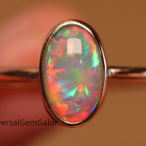 Könnte beinhalten: Ein roségoldener Ring mit einem ovalen Opal-Edelstein. Der Opal zeigt ein lebendiges Farbspiel mit roten, grünen und blauen Blitzen. Der Ring hat ein schlankes, poliertes Band, das die Brillanz des Edelsteins hervorhebt.