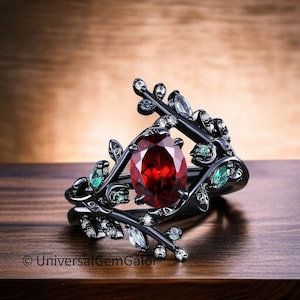 以下が含まれることがあります： 中央に大きな楕円形の赤い宝石をあしらった黒い指輪。小さな緑と透明の宝石をあしらったフローラルデザインで囲まれています。指輪は木製の表面に置かれています。