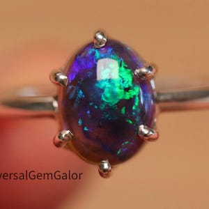 May include: A silver ring with a dark opal gemstone. The oval opal displays green, blue, and purple hues. The ring has six prongs holding the stone, and the band is polished silver. The opal's colours are vivid.