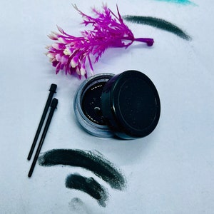 Puede incluir: Un tarro de delineador de ojos negro con una tapa negra y dos aplicadores negros. El tarro está abierto y el delineador de ojos es visible. El delineador de ojos está rodeado de manchas negras sobre un fondo blanco. Una flor morada se encuentra en la esquina superior izquierda de la imagen.
