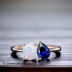 Könnte beinhalten: Ein roségoldener Ring mit einem ovalen Opal und einem tropfenförmigen Saphir. Der Opal ist weiß mit schillernden Reflexen, der Saphir tiefblau. Der Ring liegt auf einer dunklen Holzoberfläche.