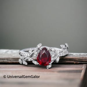May include: A pear-shaped ruby gemstone set in a silver ring with a delicate leaf design. The ring is accented with smaller diamonds.