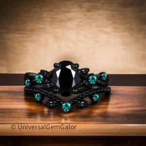 Può includere: Un set di anelli di fidanzamento neri con una pietra nera taglio ovale e pietre di accento verde acqua. L'anello ha un design floreale ed è posto su una superficie di legno.
