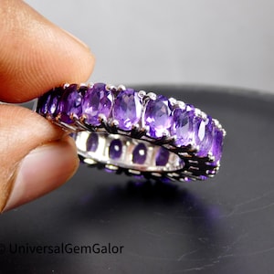 May include: A silver band ring with a row of oval-shaped amethyst gemstones. The ring is set with a total of 14 amethysts, each with a deep purple hue.