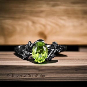 Może przedstawiać: Czarny metalowy pierścionek z dużym, owalnym, jasnozielonym kamieniem szlachetnym. Pierścionek ma kwiatowy wzór z małymi czarnymi kamieniami. Pierścionek jest umieszczony na drewnianym tle.