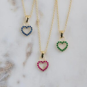 May include: Three gold necklaces with heart-shaped pendants. Each pendant is outlined with small, colorful gemstones. One pendant features blue stones, another green, and the third is red. The necklaces are displayed on a white marble surface.