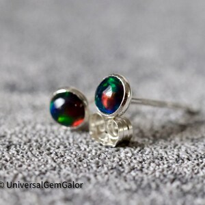 Puede incluir: Un par de pendientes de botón con ópalos negros redondos que muestran destellos de verde, rojo y azul. Los ópalos están engastados en un bisel de plata pulida. Los pendientes se muestran sobre una superficie gris texturizada.