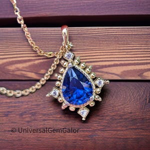 May include: A gold pendant necklace featuring a large, pear-shaped, deep blue gemstone. The pendant is surrounded by smaller white gemstones and gold accents. The necklace is displayed on a wooden surface. The text "UniversalGemGalor" is at the bottom.