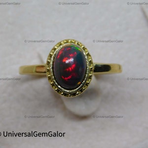 May include: A gold ring with an oval black opal displaying red and green iridescence. The opal is set in a decorative bezel. The ring's band is a simple, polished gold design.