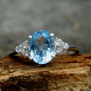 May include: A silver ring with a large oval-shaped blue topaz gemstone and smaller clear gemstones on the band.