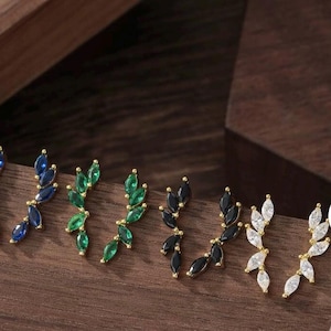May include: An assortment of leaf-shaped earrings with gold-colored posts. The earrings feature marquise-cut gemstones in various colors, including blue, green, black, and white. The earrings are displayed on a wooden surface.