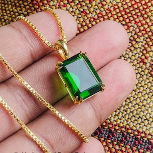May include: A gold-colored necklace featuring a rectangular, emerald-green gemstone pendant. The pendant is set in a gold frame and hangs from a delicate gold chain. The image is set against a textured background.
