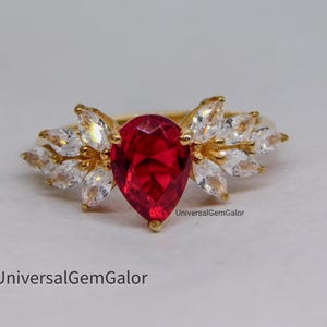 May include: A gold ring featuring a large, pear-shaped, red gemstone at the center, surrounded by smaller, white, marquise-cut stones. The ring has a delicate, floral-like design. The ring is set against a neutral background.
