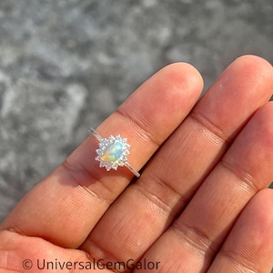 Könnte beinhalten: Ein silberner Ring mit einem ovalen Opal-Edelstein, umgeben von kleinen, klaren Edelsteinen. Der Opal zeigt blaue, gelbe und weiße Farbtöne. Der Ring sitzt auf einem zarten Band. Der Text "UniversalGemGalor" ist sichtbar.