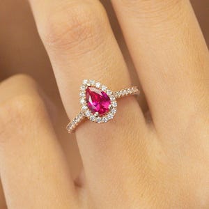 Könnte beinhalten: Ein roségoldener Ring mit einem tropfenförmigen, leuchtend rosa Edelstein, umgeben von einem Kranz aus kleinen, klaren Steinen. Das Band ist ebenfalls mit kleinen, klaren Steinen besetzt. Schmuck.