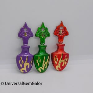 Pode incluir: Três rolhas de garrafa decorativas em forma de garrafa com uma parte superior pontiaguda. As rolhas de garrafa são roxas, verdes e vermelhas com detalhes e desenhos dourados.